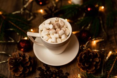 Noel ışıklarıyla dolu bir masada beyaz bir fincanda marşmelov. Noel ağacı ve koniler masanın üstünde. Yüksek kalite fotoğraf