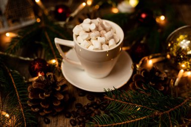 Noel ışıklarıyla dolu bir masada beyaz bir fincanda marşmelov. Noel ağacı ve koniler masanın üstünde. Yüksek kalite fotoğraf