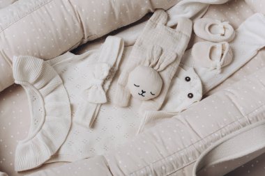 Moda, yeni doğan esasları, ıvır zıvır ve aksesuarlar. Bebek takımları, kız şamatası, tozluklar, aksesuarlar, baş yayı, botlar, bej kozanın üstündeki tavşan oyuncağı, yuva arka planı. Üst görünüm.