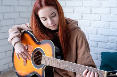 young caucasian red-haired woman sitting on rug and playing acoustic guitar at home