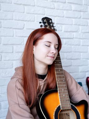 young caucasian red-haired woman sitting on rug and playing acoustic guitar at home