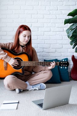 young caucasian red-haired woman learning to play acoustic guitar at home, making notes and using laptop