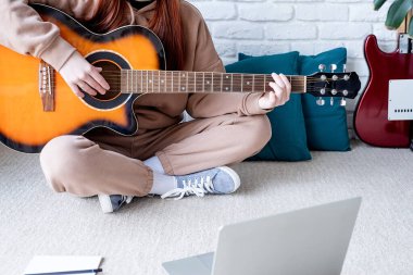 young caucasian red-haired woman learning to play acoustic guitar at home, making notes and using laptop