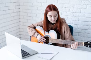 young caucasian red-haired woman learning to play acoustic guitar at home, making notes and using laptop
