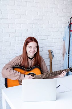 young caucasian red-haired woman learning to play acoustic guitar at home, making notes and using laptop