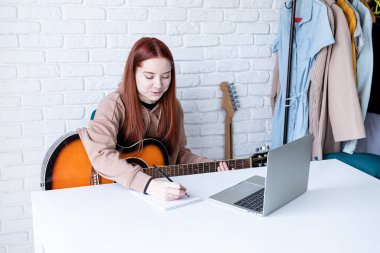 young caucasian red-haired woman learning to play acoustic guitar at home, making notes and using laptop