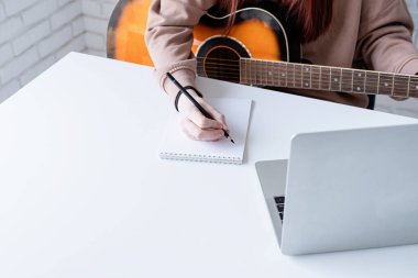 young caucasian red-haired woman learning to play acoustic guitar at home, making notes and using laptop