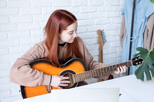 young caucasian red-haired woman learning to play acoustic guitar at home, making notes and using laptop