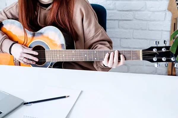 young caucasian red-haired woman learning to play acoustic guitar at home, making notes and using laptop