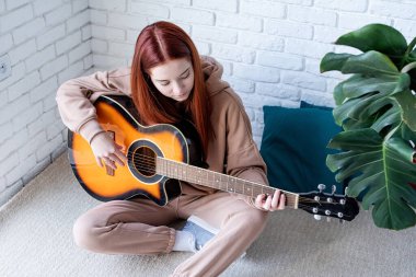 young caucasian red-haired woman sitting on rug and playing acoustic guitar at home