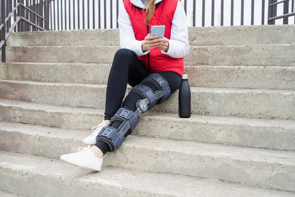 Woman wearing knee brace or orthosis after leg surgery, sitting on stairs ourdoors. Medical and healthcare concept.