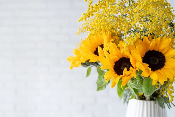 Blossom of Mimosa flowers and sunflowers in white ceramic vase on white brick wall background with space for text. Spring concept, yellow color