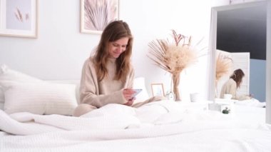 Evde yatakta oturan, kahvaltı yapan ve akıllı telefon kullanan bir kadın. Genç milenyum kadını yatak odasında sabah rutinini yapıyor.