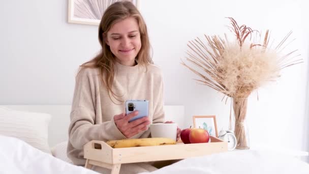 Kobieta Siedząca Domu Łóżku Jedząca Śniadanie Używająca Smartfona Młoda Tysiąclecia — Wideo stockowe