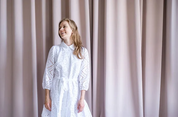 ベージュのカーテンの背景に立って白い夏のファッションドレスで美しい白人女性 離れて見て パステルカラー — ストック写真