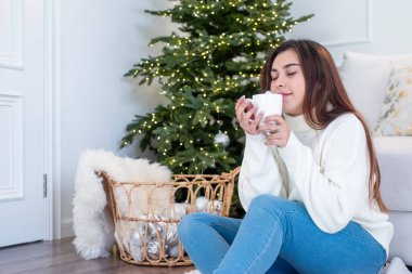 Mutlu Noeller ve mutlu yıllar. Sıcak beyaz kazaklı bir kadın Noel arifesinde evinin penceresinde oturmuş elinde lokum, arkasında köknar ağacı olan bir bardak tutuyordu.