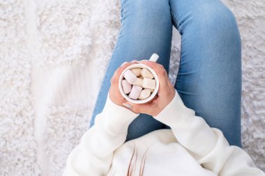 Merry Christmas and Happy New Year. Woman in warm white winter sweater holding cup with marshmallows, fir tree behind