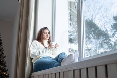 Merry Christmas and Happy New Year. Woman in warm white winter sweater sitting on the window at home at christmas eve holding cup with marshmallows, fir tree behind