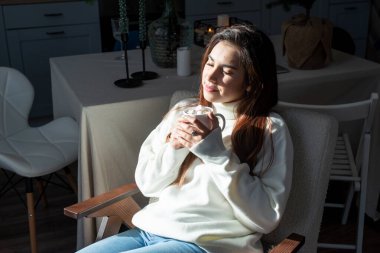 Merry Christmas and Happy New Year. Woman in warm white winter sweater sitting on the window at home at christmas eve holding cup with marshmallows, fir tree behind