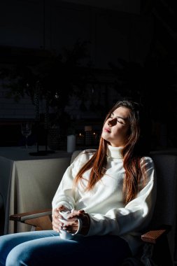 Merry Christmas and Happy New Year. Woman in warm white winter sweater sitting on the window at home at christmas eve holding cup with marshmallows, fir tree behind
