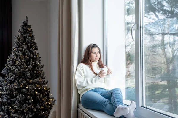 メリークリスマスとハッピーニューイヤー 暖かい白い冬のセーターで女性は家の窓の上に座っていますクリスマスの前夜にカップを保持します — ストック写真