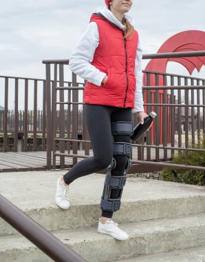 Woman wearing sport clothes and knee brace or orthosis after leg surgery, walking down the stairs in the park. Medical and healthcare concept.