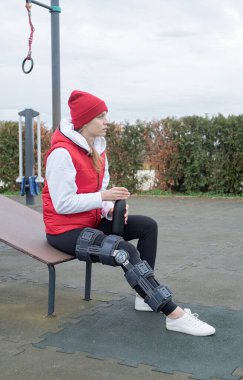 Woman wearing sport clothes and knee brace or orthosis after leg surgery, walking down the stairs in the park. Medical and healthcare concept.