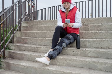 Woman wearing sport clothes and knee brace or orthosis after leg surgery, walking down the stairs in the park. Medical and healthcare concept.