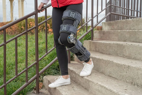 Woman wearing sport clothes and knee brace or orthosis after leg surgery, walking down the stairs in the park. Medical and healthcare concept.