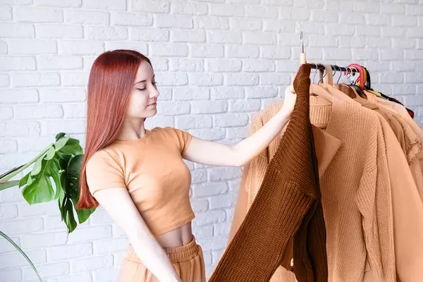 2024年的颜色 Peach Fuzz 穿着米色外套的年轻漂亮的女人站在衣架前挑选服装 选择衣橱 造型师 购物者 服装设计 图库图片