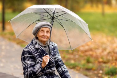 Gri ceketli ve bereli yaşlı bir kadın sonbaharda elinde şeffaf bir şemsiye olan bir bankta oturur ve doğrudan kameraya bakar. Huzurlu ve ılık bir sonbahar havası..