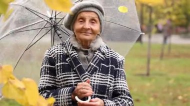 80 yaşından büyük bir kadın, gülümsüyor ve şeffaf bir şemsiyeyi açık havada çeviriyor. Sarı sonbahar yapraklarıyla çevriliyken kameraya bakar. Doğada yürek ısıtan bir sevinç anı.
