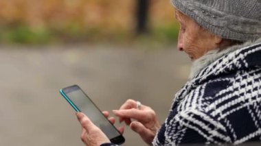 Yaşlı bir kadın yağmurlu bir sonbahar parkında elinde akıllı bir telefonla duruyor. Ekrana vuruyor ve bir şey seçmeye odaklanıyor. Sahne sakin ve yansıtıcı..