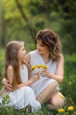 Beyaz elbiseli bir kadın ve genç bir kız çayırda yeşil çimlerde oturuyor, ellerinde sarı karahindiba buketleri, güneşli bir bahar gününde gülümsüyor ve birbirlerine bakıyorlar..