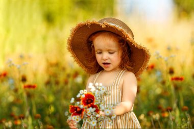 Sarı çizgili elbisesi ve geniş hasır şapkası olan tatlı kızıl saçlı kız elinde bir buket kır çiçeğiyle çiçek açmış bir çayırda duruyor..