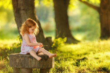 Sevimli kızıl saçlı, pembe yaz elbisesi içinde küçük bir kız. Güneşli bir ormanda, tahta bir kütüğün üzerinde çıplak ayakla oturuyor, elinde küçük bir çiçek tutuyor ve düşünceli bir şekilde aşağıya bakıyor.. 