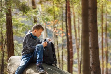 Genç bir çocuk ormanda büyük bir kayanın üzerinde oturuyor, sırt çantasını açıyor. Günlük kıyafetlerle gezen çocuk doğa yürüyüşü, keşif ve çocukluk macerası kavramı..
