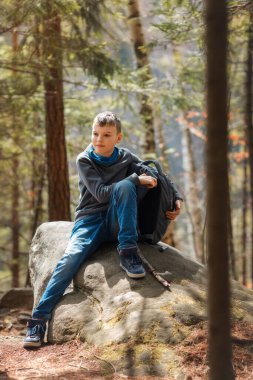 Bir çocuk yürüyüş sırasında ormanda büyük bir kayanın üzerinde oturuyor, günlük kıyafetler giyiyor ve bir sırt çantası tutuyor. Güneş ışığı ağaçların arasından süzülüp, dışarıda huzurlu bir atmosfer yaratıyor.. 