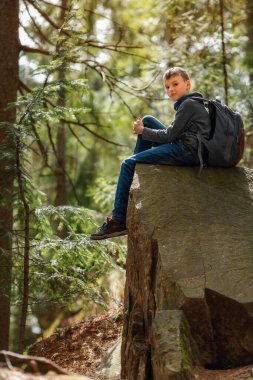 Bir çocuk yürüyüş sırasında ormanda büyük bir kayanın üzerinde oturuyor, günlük kıyafetler giyiyor ve bir sırt çantası tutuyor. Güneş ışığı ağaçların arasından süzülüp, dışarıda huzurlu bir atmosfer yaratıyor.. 