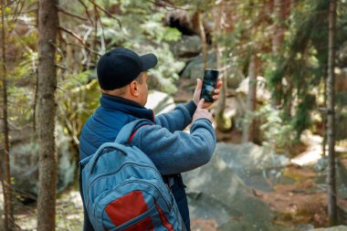 Sırt çantalı bir adam, akıllı telefonlu orman manzarasının fotoğrafını ya da videosunu çekiyor. Yürüyüşçü doğa yürüyüşü sırasında anları yakalıyor.