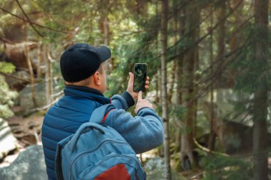 Sırt çantalı bir adam, akıllı telefonlu orman manzarasının fotoğrafını ya da videosunu çekiyor. Yürüyüşçü doğa yürüyüşü sırasında anları yakalıyor.