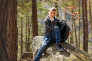Bir çocuk yürüyüş sırasında ormanda büyük bir kayanın üzerinde oturuyor, günlük kıyafetler giyiyor ve bir sırt çantası tutuyor. Güneş ışığı ağaçların arasından süzülüp, dışarıda huzurlu bir atmosfer yaratıyor.. 