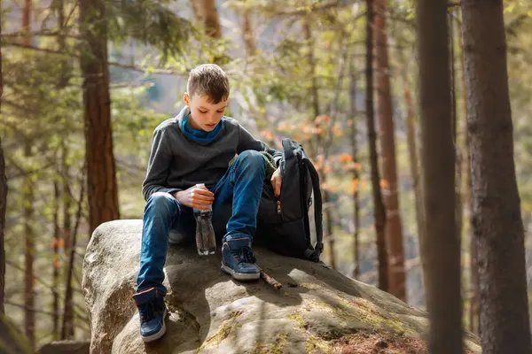 Ormanın ortasında, yosun kaplı bir kayanın üzerinde elinde su şişesiyle oturan genç bir yürüyüşçü. Ağaç gövdeleri arka planda görünür ve güneş ışığı yaprakların arasından süzülür..