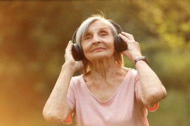 Yaşlı kadın kablosuz kulaklıkla müzik dinliyor, doğada huzur içinde duruyor ve nazikçe gülümsüyor..