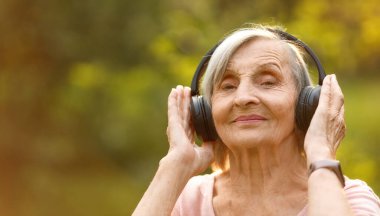 Yaşlı, gri saçlı, kulaklıklı, neşeli gülümseyen, güneşli bir bahçede dikilen bir kadın..