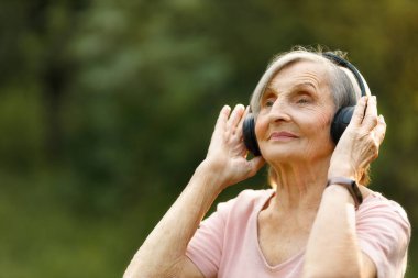 Yaşlı kadın kablosuz kulaklıkla müzik dinliyor, doğada huzur içinde duruyor ve nazikçe gülümsüyor..