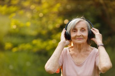 Yaşlı gri saçlı kadın, gözleri kapalı, kablosuz kulaklıkları nazikçe elinde, müziğe dalmış bir şekilde dışarıda duruyor..