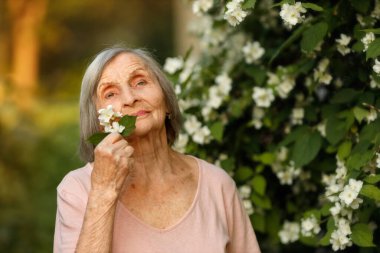 Yasemin çalılarının yanında duran yaşlı bir kadın, yanağına hafifçe leylak dalı bastırıyor ve uzaklığa bakıyor..