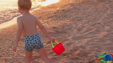 Üç yaşında bir çocuk gün batımında denize koşup kırmızı kovaya su dolduruyor. Sahil kenarında sıcak altın bir ışık, tasasız bir çocukluk anı.