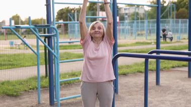 80 yaşın üzerinde yaşlı bir kadın dışarıda nefes alma egzersizi yapıyor. Kollarını derin bir nefes alarak kaldırıyor ve onları nefes vererek, denge ve dinginliği göstererek aşağı indiriyor..